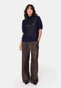 Tengerészkék, textúrázott hálós blúz puffos ujjakkal és magas nyakkivágással, magas derekú, barna, plisszírozott nadrággal párosítva.