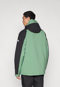 Χειμερινό μπουφάν σκι με πράσινο σώμα και μαύρα μανίκια, με κουκούλα, υφή στο ύφασμα και λογότυπο στον αριστερό ώμο.