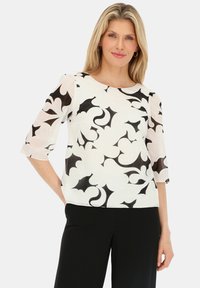 Biała bluzka z czarnym florystycznym wzorem, półprzezroczystymi rękawami, okrągłym dekoltem oraz luźnym krojem, w połączeniu z czarnymi spodniami.