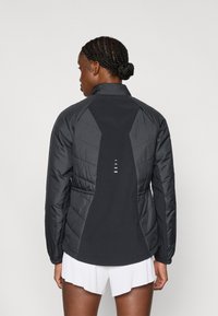 Μαύρο μπουφάν τύπου puffer με υφή κουμπότρυπα, ψηλό γιακά και ανακλαστικές λεπτομέρειες. Διαθέτει σφηνοειδή μέση και κομψό σχέδιο.