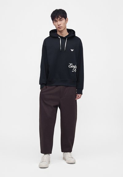 Jeune homme portant un sweat à capuche noir avec des cordons blancs, un pantalon ample foncé et des baskets blanches, debout devant un fond blanc uni.