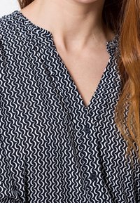 Blusa blu navy con un motivo a onde bianche, con scollatura a v e dettaglio dei bottoni. Il tessuto sembra leggero con una texture morbida.