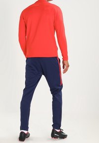 Punainen pitkähiha, joka on yhdistetty tummansinisiin urheiluhousuihin, joissa on punaiset sivuraidat. Housuissa on vetoketjut nilkoissa ja tekstuuripinta.