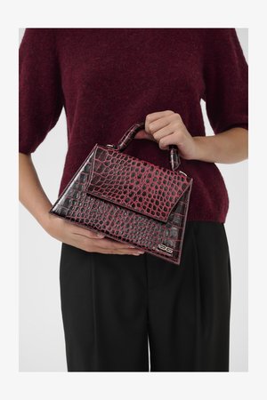 Personne tenant un petit sac à main maroon et noir à motif crocodile avec une anse, portant un pull bordeaux et un pantalon noir.