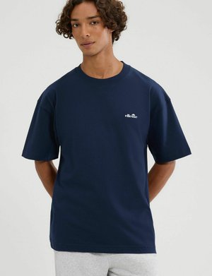 Joven con cabello rizado vistiendo una camiseta azul marino de ellesse y pantalones gris claro, de pie con las manos detrás de la espalda.