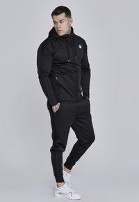 Czarny dres sportowy wykonany z gładkiego materiału, z kapturem zapinanym na zamek i logo oraz bocznymi kieszeniami, w połączeniu z zwężającymi się spodniami i białymi sneakersami.
