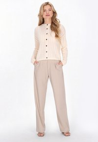 Crèmewitte gebreide cardigan met een gestructureerd patroon en zes knopen, gecombineerd met lichtbeige wijde broekspijpen en open-toe sandalen.