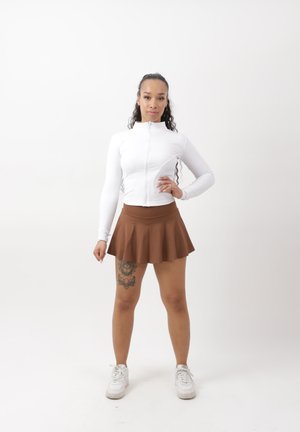 Frau steht mit der Hand auf der Hüfte, trägt eine weiße Jacke mit Reißverschluss, einen braunen Rock, weiße Sneaker und ein großes Tattoo am Oberschenkel.