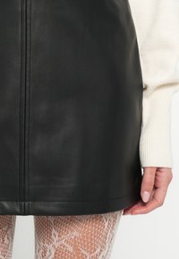 Čierna kožená mini sukňa s hladkou textúrou, s detailom švu. Kombinovaná so svetlým pleteným rukávom a sieťovanými pančuchami.