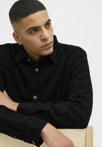 Camisa negra de textura hecha de tela tejida con cuello, presenta botones de madera y mangas largas, con los brazos descansando sobre una superficie de madera clara.