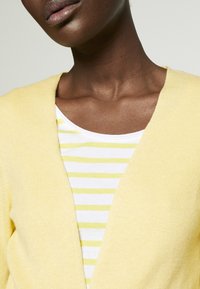 Gul strikket cardigan med V-hals design, lagd over en hvid skjorte med tynde gule striber. Glat tekstur og afslappet pasform.