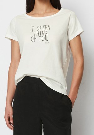 Vrouw die een wit T-shirt met zwarte tekst "I OFTEN THINK OF YOU. FUTURE" en een zwarte broek draagt, één hand in de zak, staand tegen een neutrale achtergrond.