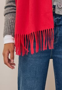 Écharpe rouge en tissu doux avec bords frangés, portée sur un pull en maille gris, associée à un jean en denim bleu.