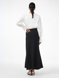 Blouse blanche avec des accents en broderie sur les manches, associée à une jupe longue et fluide noire. Le modèle se tient de dos face à l'appareil photo, mettant en évidence une silhouette élégante.
