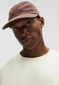 Hnedá baseballová čiapka vyrobená z textílie s zakriveným šiltom, s embosovaným logom vpredu. Nosená s svetlým crew neck mikinou.