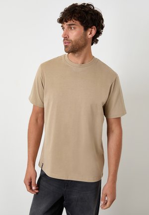 Uomo con capelli ricci che indossa una maglietta beige a maniche corte e jeans neri, guarda verso sinistra su uno sfondo chiaro uniforme.