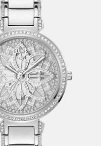 Zilveren Guess horloge met een gedetailleerde bloemenontwerp wijzerplaat, omringd door kristallen, en een gepolijst metalen band met glanzende accenten.