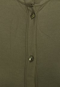 Camisa abotoada em verde oliva, com um tecido texturizado, cinco botões na frente e detalhes de costura subtil.