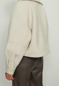 Kremowa, teksturowana kurtka z wyraźnym kołnierzem, luźnym krojem i zapinanymi na guziki mankietami, w połączeniu z brązowymi skórzanymi spodniami.