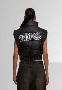Black padded vest with a high collar and cropped design. Features large white "INGRS" lettering on the back, smooth texture, and shiny finish.
