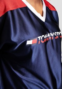 Camiseta deportiva azul marino con mangas acentuadas en rojo, cuello en V y cuello blanco. "TOMMY SPORT" impreso en el frente con detalle de bloques de color horizontales.