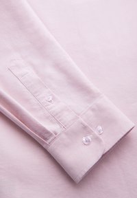 Poignet de chemise à manches longues rose clair avec deux boutons transparents et coutures détaillées sur un tissu lisse.