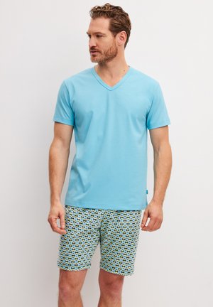 Uomo con indosso una maglietta a V azzurro chiaro e pantaloncini con motivo a disegni, in piedi e che guarda verso sinistra su sfondo bianco semplice.