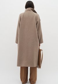 Cappotto di lana beige con colletto alto, maniche lunghe e taglio dritto, indossato sopra pantaloni marroni, con una borsa a tracolla marrone chiaro.