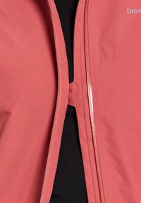 Koraalkleurige jas met een gladde textuur, voorzien van een centrale ritssluiting en een knoopsluiting. Zwart binnenvak is zichtbaar eronder.