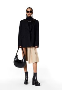 Blazer noir à double boutonnage sur un col roulé noir, jupe plissée beige, bottes à talons larges noires et sac hobo noir.