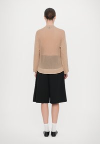 Cardigan lavorato a maglia beige con maniche lunghe, pannelli laterali testurizzati e un design leggero. Abbinato a pantaloni corti neri до al ginocchio e calzini.