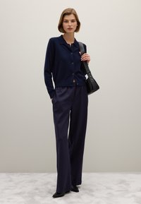 Marineblauwe cardigan met knopen, gecombineerd met losse marineblauwe broek en zwarte schoenen. De look is accessorized met een kleine zwarte crossbody tas.