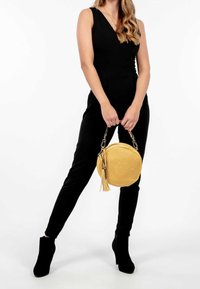 Vrouw in zwarte mouwloze jumpsuit en zwarte enkellaarzen, die een ronde gele handtas met een franjes vasthoudt, staande tegen een witte achtergrond.