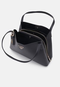 Sac à main noir avec deux poignées, un compartiment zippé et un logo doré visible ainsi qu'une étiquette de marque sur fond blanc.