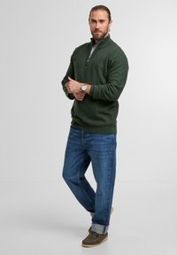 Grüne Zip-Up-Pullover aus weichem Stoff, kombiniert mit blauen Jeans und braunen Schuhen. Der Look zeichnet sich durch einen lässigen Stil mit hochgekrempelten Ärmeln und einer Uhr aus.