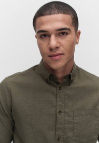 Olijfgroene button-up shirt van textuurstof, met een kraag, voorzak en bruine knopen. Close-upportret van een jonge man.
