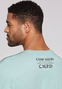 Světle modré tričko s krátkým rukávem a kulatým výstřihem, s černým potiskem na zadní straně a jemnou strukturou látky.