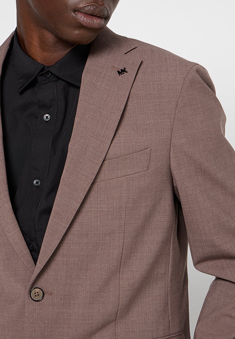 Blazer marrone con texture, chiusura a un bottone e taschino sul lato sinistro del petto, abbinato a una camicia nera con dettagli sui bottoni.