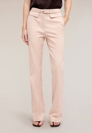 Mujer que lleva pantalones anchos de tiro alto de color rosa claro con bolsillos, un cinturón, sandalias negras de punta abierta y pulseras doradas en ambas muñecas.