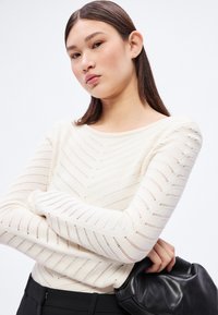 Hvit strikket genser med chevron-mønster, lange ermer og vid hals. Kombinert med en svart clutchveske med glatt tekstur.