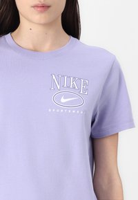 Violet medvilninis marškinėliai su apvaliu kaklu, ant kurių baltu šriftu parašyta "NIKE" ir "SPORTSWEAR", o žemiau yra ovalus Nike logotipas. Paprastas dizainas, trumpomis rankovėmis.