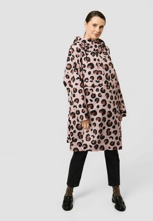 Femme portant un imperméable rose à capuche imprimé léopard, pantalon court noir, chaussettes assorties imprimées léopard et mocassins noirs, debout avec un léger sourire.