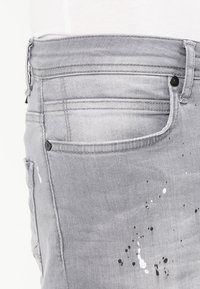 Grå denimjeans har en åtsittande silhuett, lätt slitningar och splattereffekter i svart och vitt på framsidan.