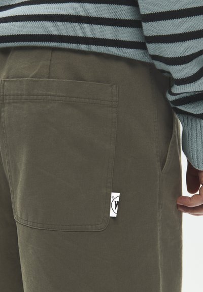 Gros plan d'une personne portant un pantalon olive avec une poche arrière carrée et une manche de pull rayée bleu et noir.