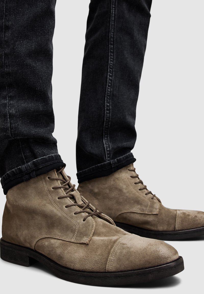 Stivaletti in suede marrone alla caviglia con punta tonda, lacci, e suola in gomma scura a contrasto, abbinati a jeans neri arrotolati ai polsini.