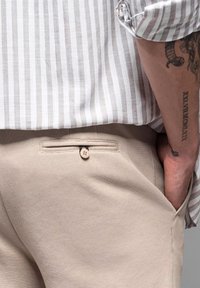 Pantalones beige texturizados con un bolsillo trasero que tiene un cierre de botón. El usuario tiene la mano en el bolsillo, llevando una camisa a rayas arriba.