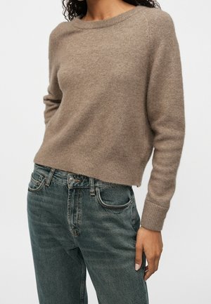 Personne portant un pull à manches longues taupe et un jean bleu délavé taille haute, debout devant un fond clair uni.