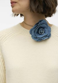 Spilla di fiori in denim blu, con petali sovrapposti, attaccata a un maglione di colore crema con scollo a costine, creando un contrasto di texture.