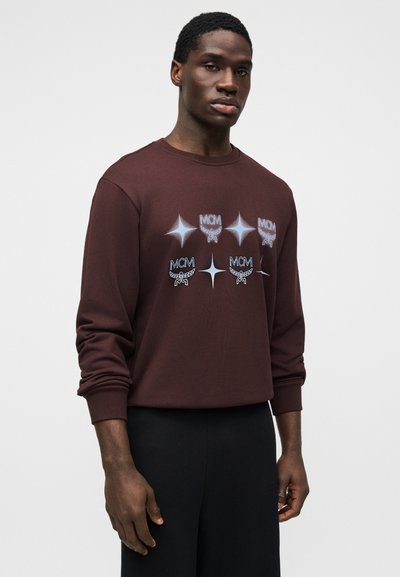 Καφέ φούτερ με λογότυπα MCM και γραφικά αστεριών, μακριά μανίκια, στρογγυλή λαιμόκοψη, κατασκευασμένο από μαλακό ύφασμα, συνδυασμένο με μαύρο παντελόνι.