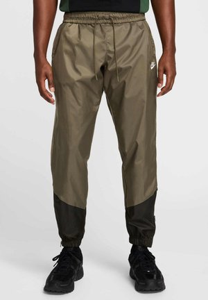 Nike track pants in olive green with black accents. Features an elastic waistband, side pockets, and zippered cuffs for adjustable fit.